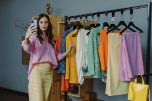 Mulher sorrindo, criadora de conteúdo de Donos de Achadinhos e-commerce, gravando um vídeo com o celular, com roupas coloridas (amarelo, laranja) em uma arara de fundo.